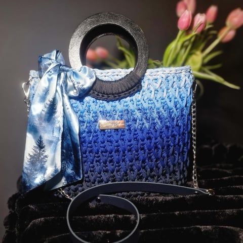 Blue textured handbag with circular gray handle, silk scarf, and fresh pink flowers