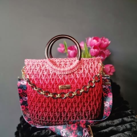 Pink textured handbag with circular metal handle, decorated with gold chain, fresh pink flowers, and green stems arranged on top
