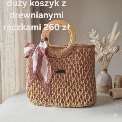 Beige woven basket bag with wooden handles, pink ribbon bow, displayed with dried flowers in vase on white surface