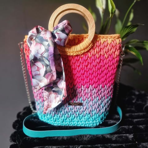 Colorful knitted bucket bag with ombre gradient from pink to turquoise, wooden handles, floral fabric bow, and turquoise shoulder strap