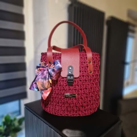 Red quilted handbag with leather handles and decorative fabric bow displayed on black pedestal
